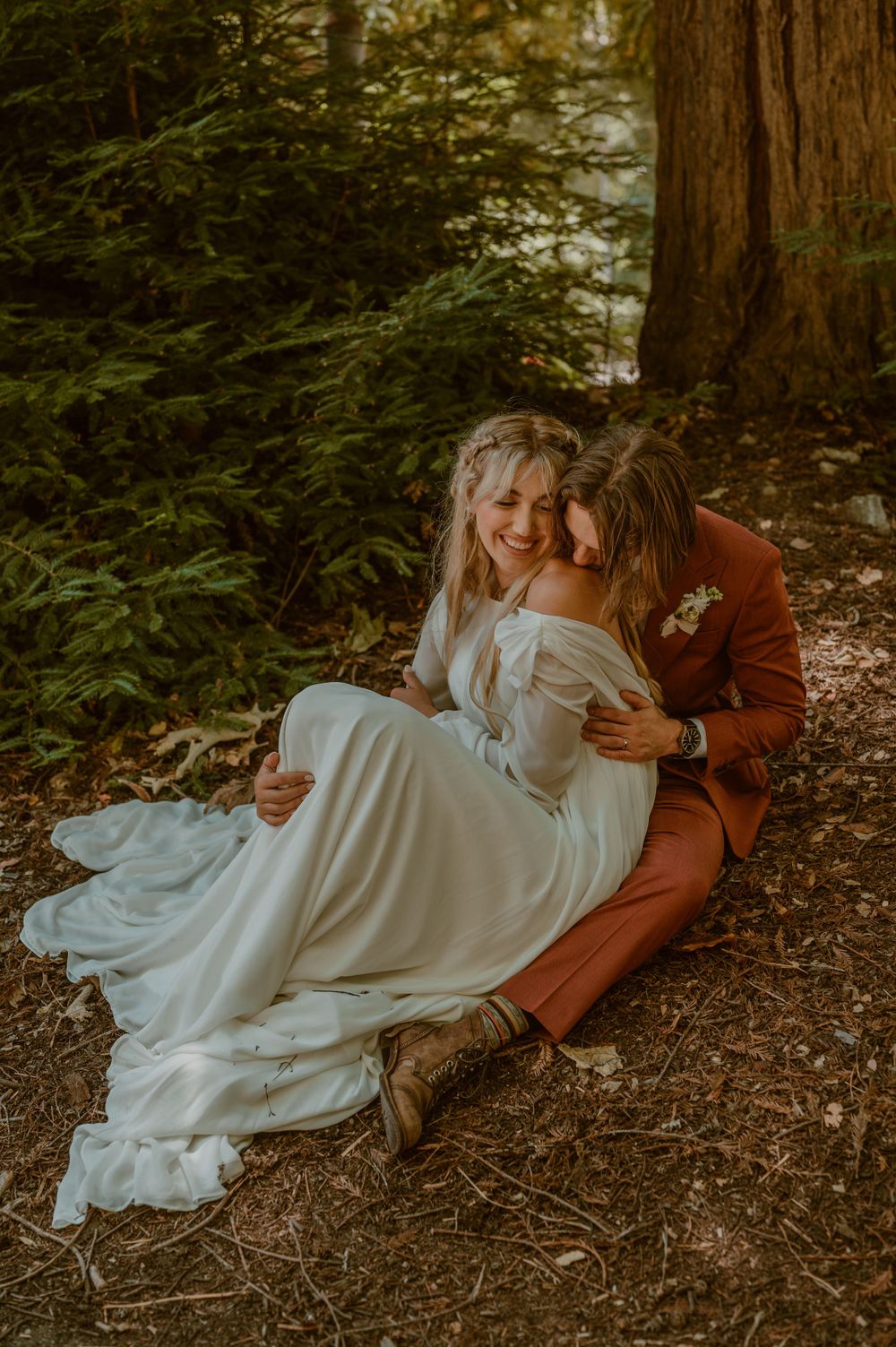 A redwoods forest wedding photo by samantha Campbell imagery shows a couple in white dress and rust colored suit sitting together on ground.