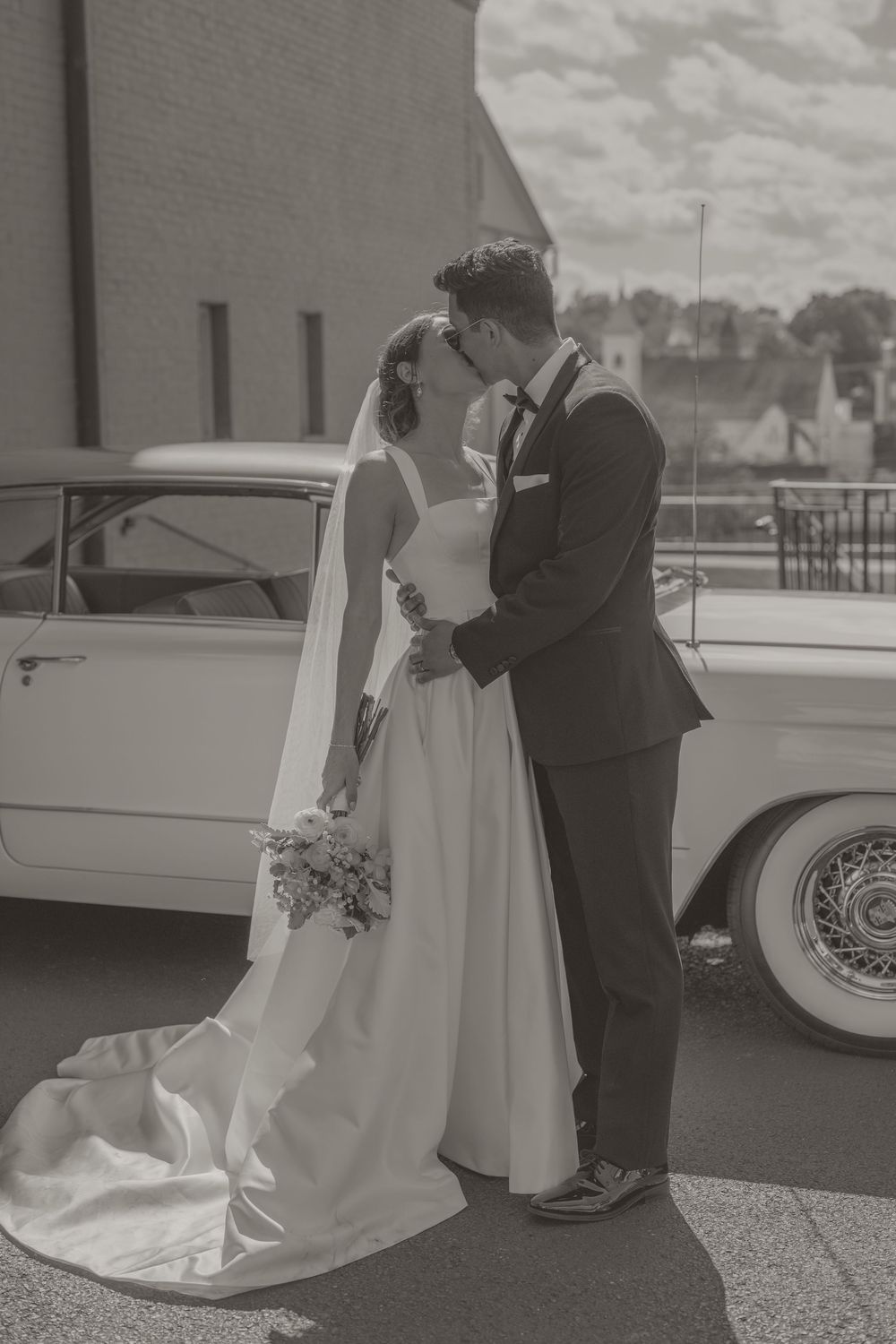 Black and white photo of a bride and groom sharing a kiss beside a vintage white car on their wedding day.