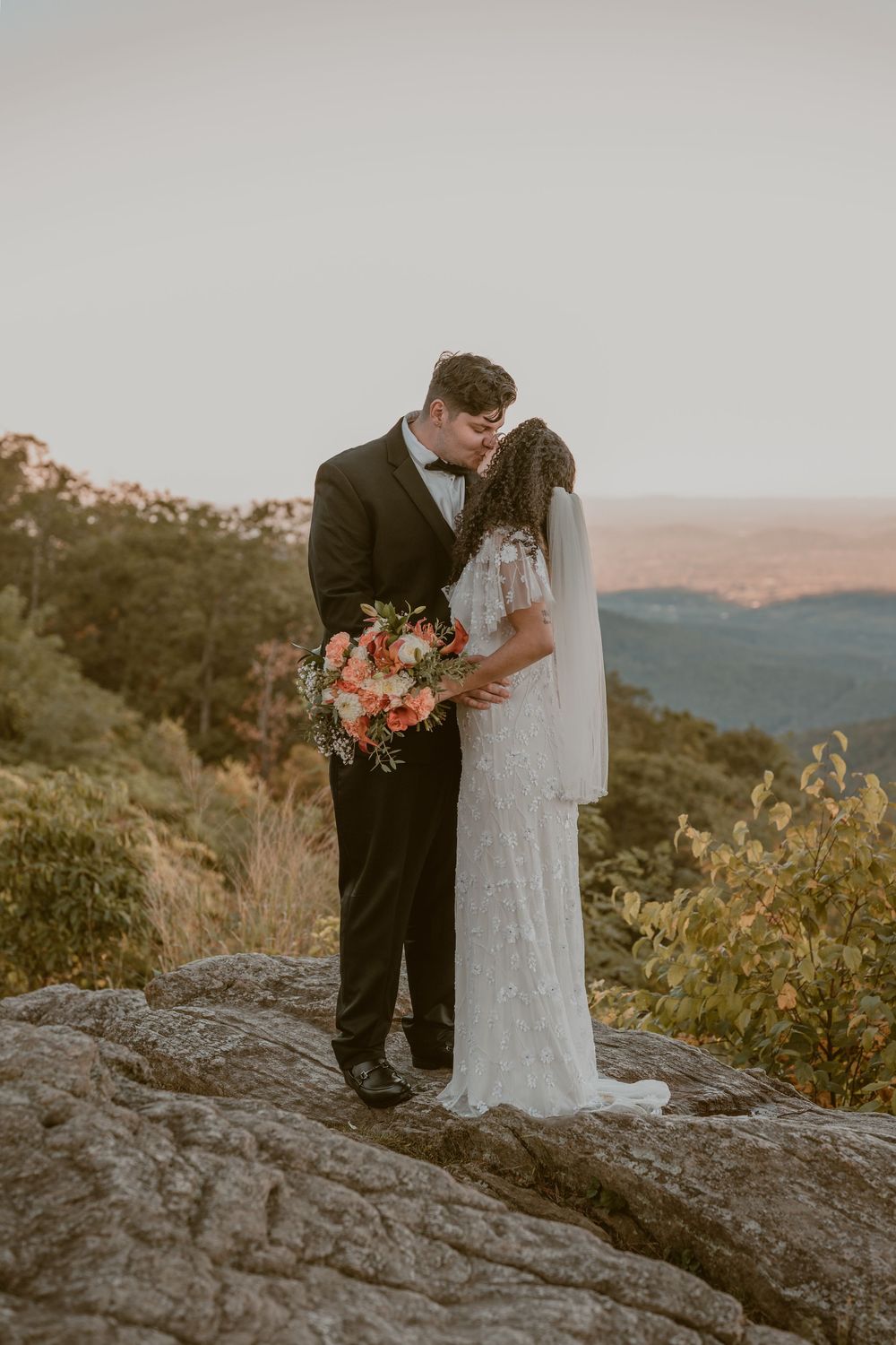 Bride and groom kissing on rocky mountain overlook at sunset, bride in lace gown holding coral bouquet.