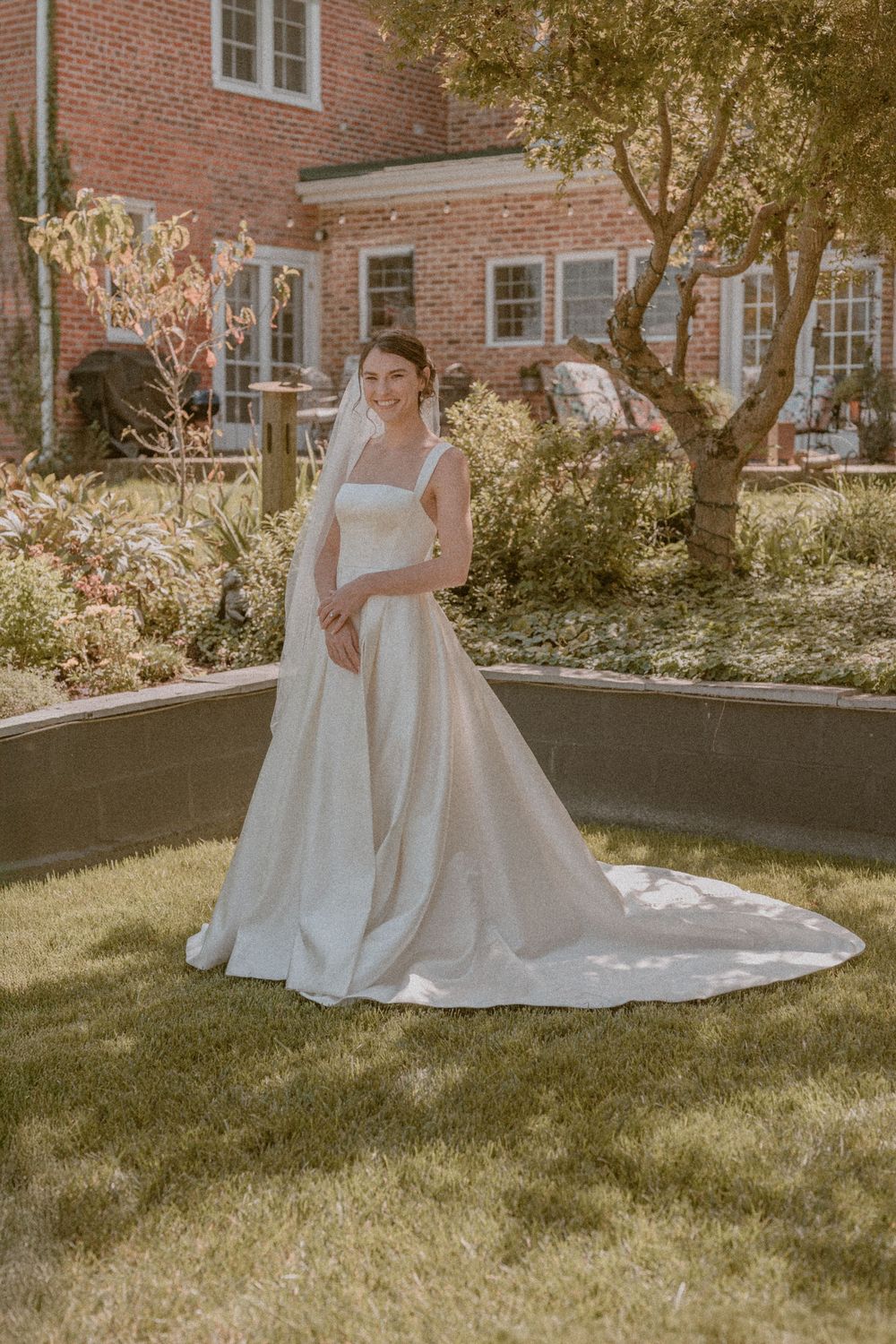 Bride in elegant white ball gown with long train and veil poses in lush garden outside brick estate.