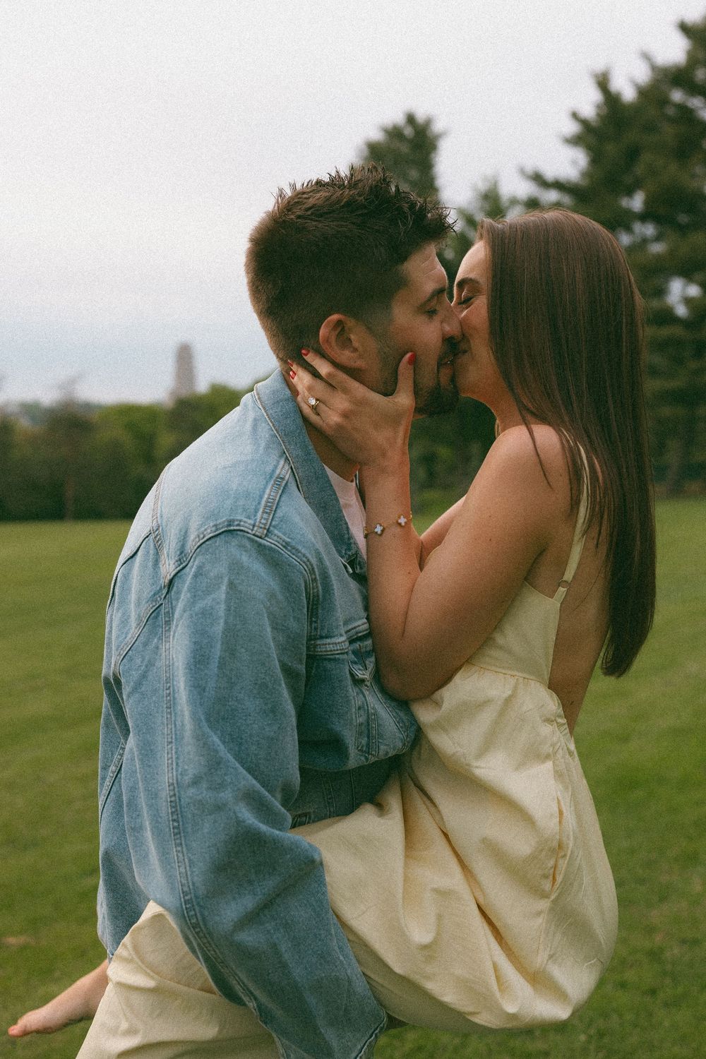 Couple sharing a romantic kiss outdoors on green grass, man in denim jacket lifting woman in cream dress.
