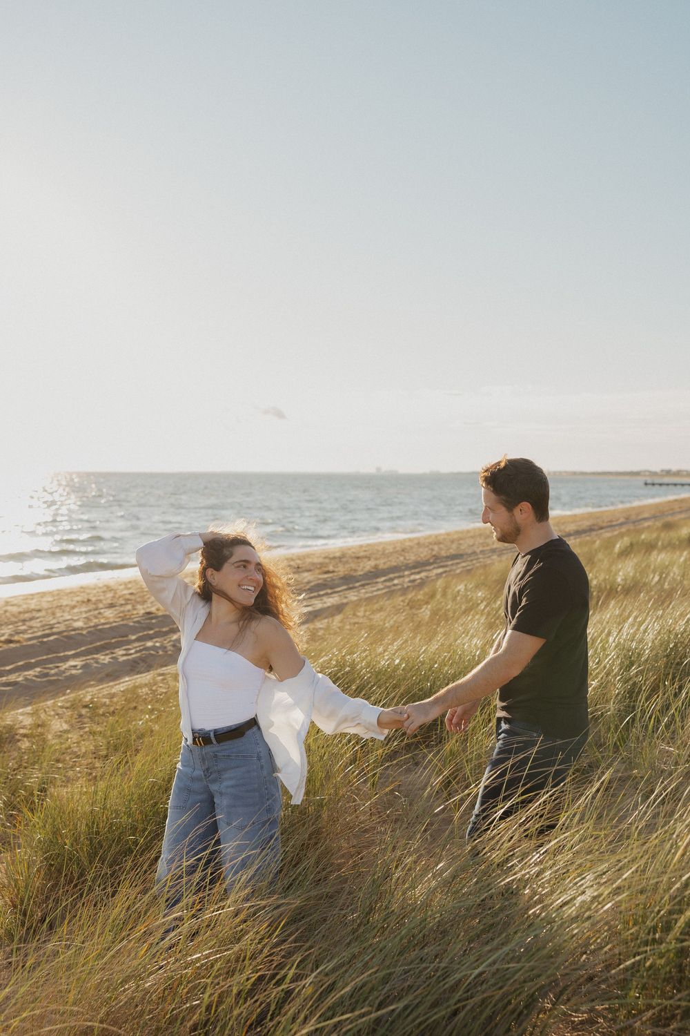 Two people sitting together on grassy dunes at sunset by the ocean with the sea breeze blowing through beach grass.