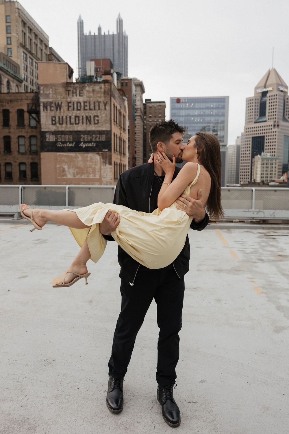 A couple shares a romantic rooftop kiss in Pittsburgh, man lifting woman in yellow dress with city skyline behind them.