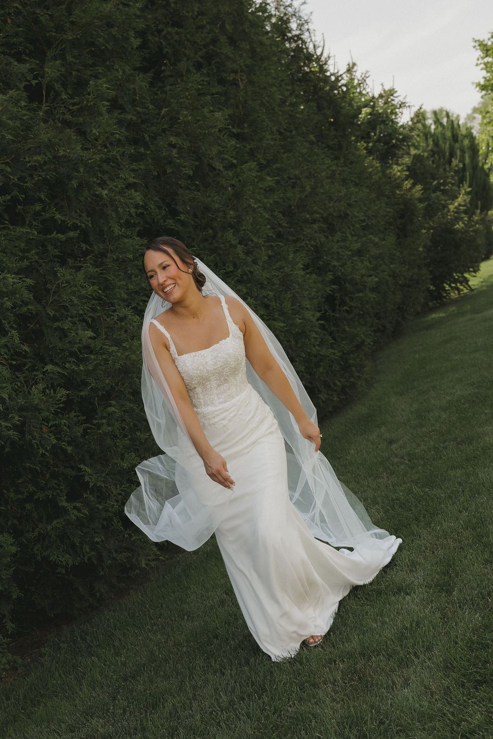 Ethereal bride in flowing white dress twirls gracefully on green grass against a backdrop of lush hedges.