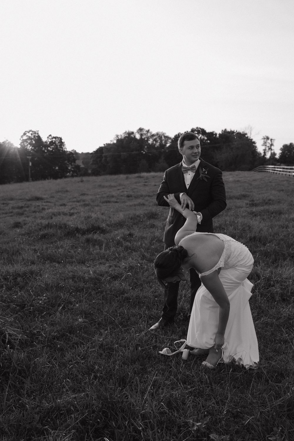 A couple in formal attire embraces romantically in a grassy field at sunset.