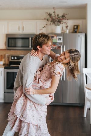 A romantic couple dancing together in their kitchen, with her wearing a pink ruffled dress and him in a beige sweater.