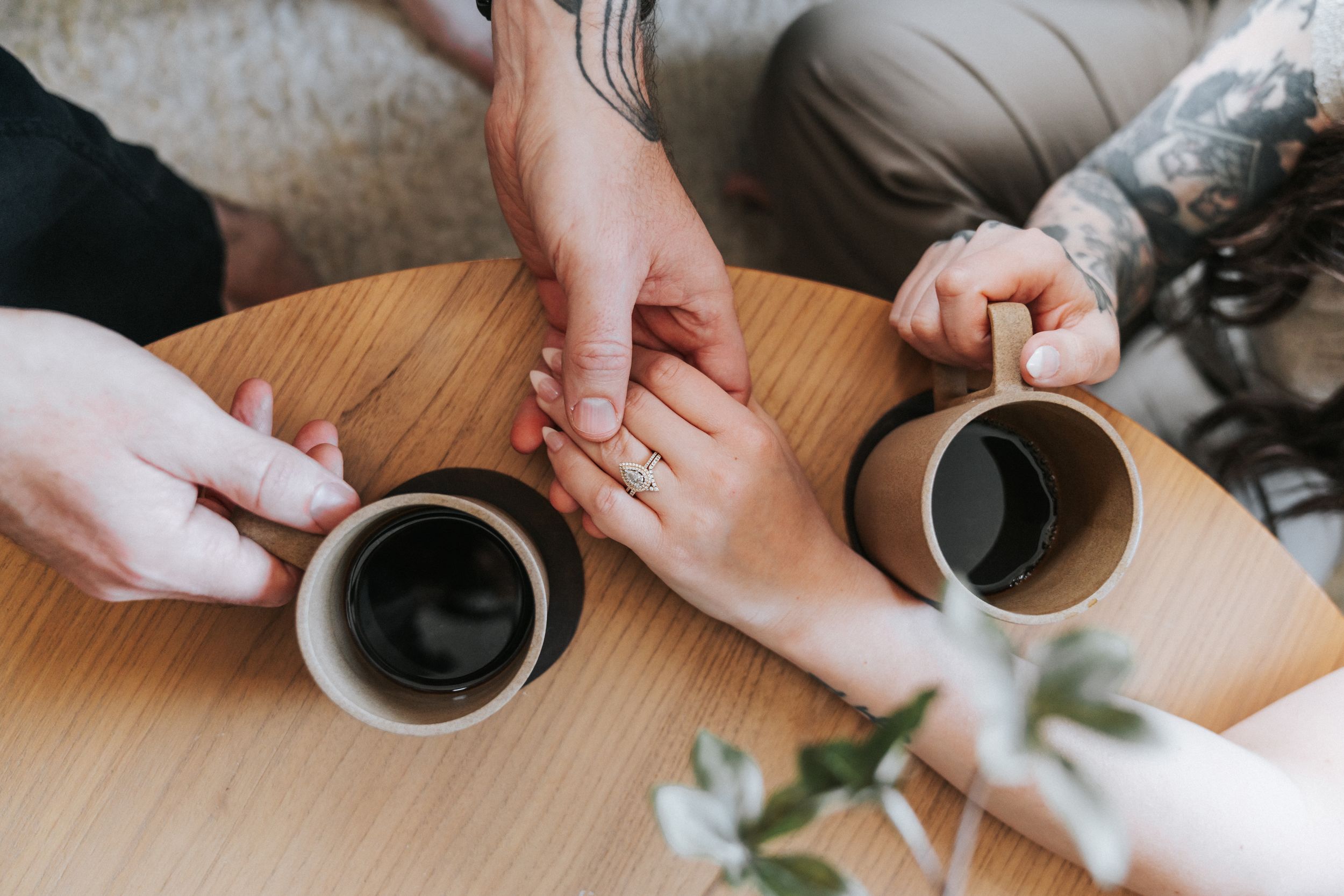 Two people in casual attire hold coffee cups while sharing an intimate moment at a wooden table.