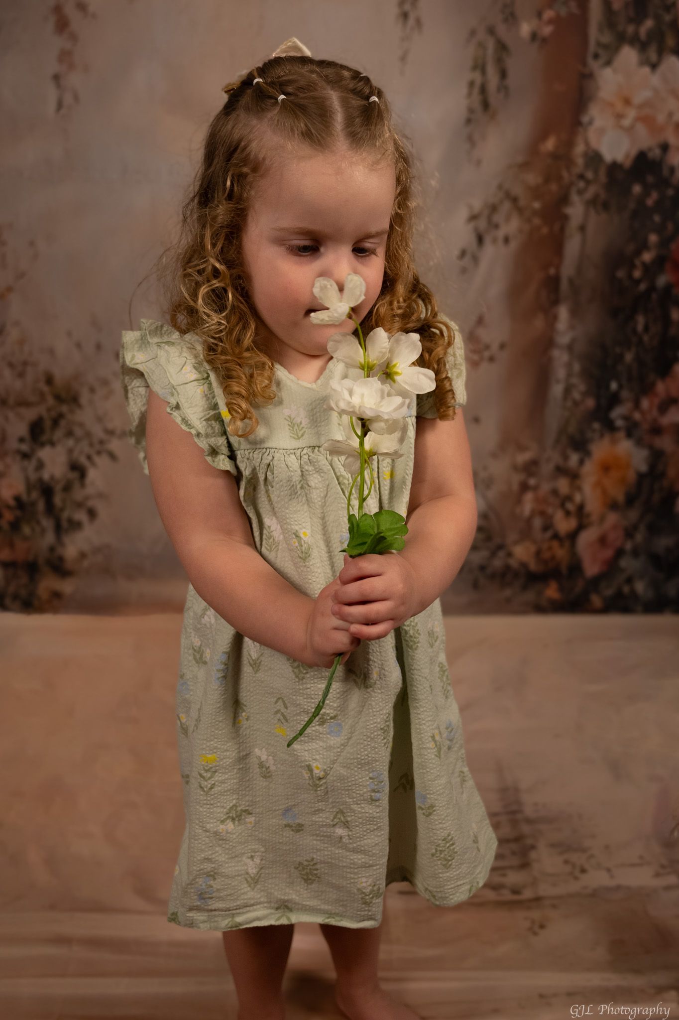 A young girl in a sage green floral dress holds white flowers while standing before a floral backdrop.