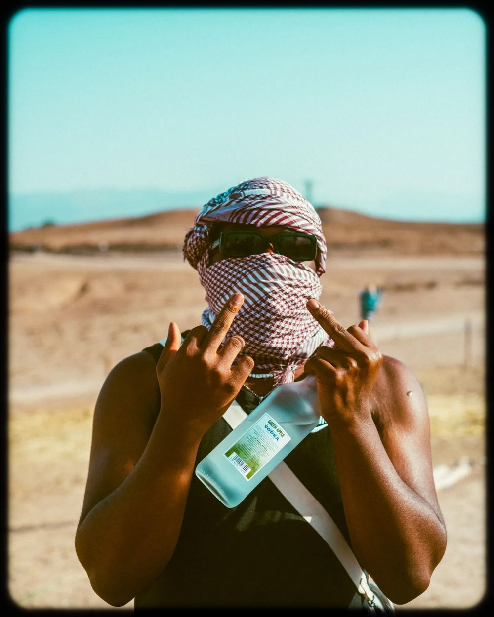 Person wearing a striped headscarf covers their face while holding a phone in a desert landscape.