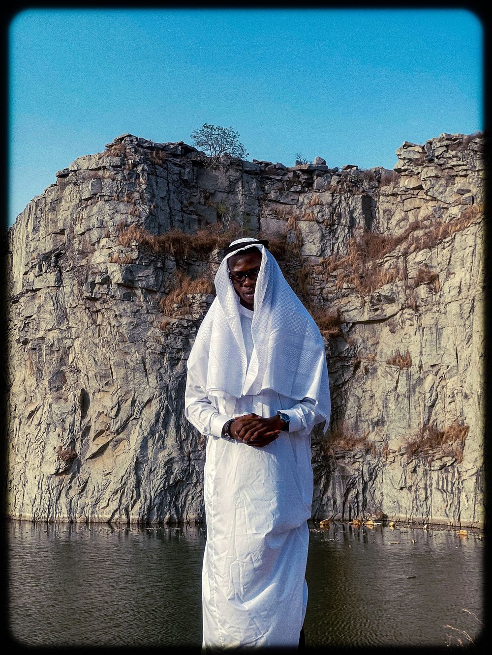 A person in white clothing stands against a dramatic rocky cliff face overlooking a serene body of water.