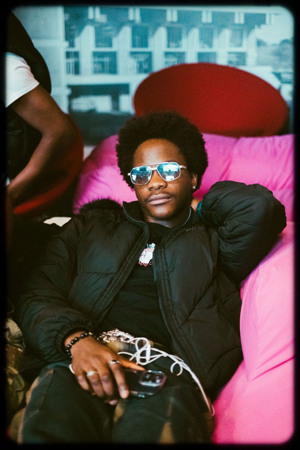 Stylish person in black puffer jacket and reflective sunglasses relaxes on pink chair in retro film photograph.