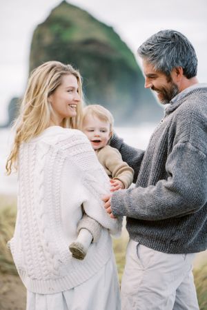 A family shares an intimate moment at the beach with dramatic coastal rock formations behind them.