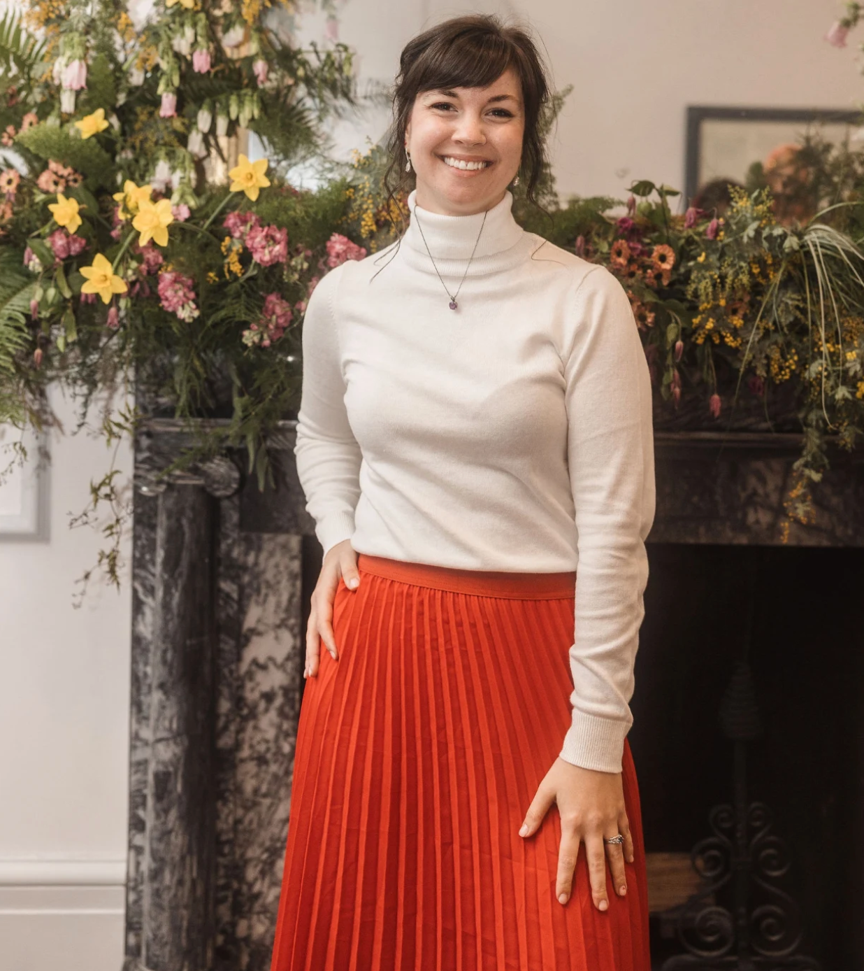 Person wearing a white turtleneck and bright red pleated skirt standing by floral arrangements.