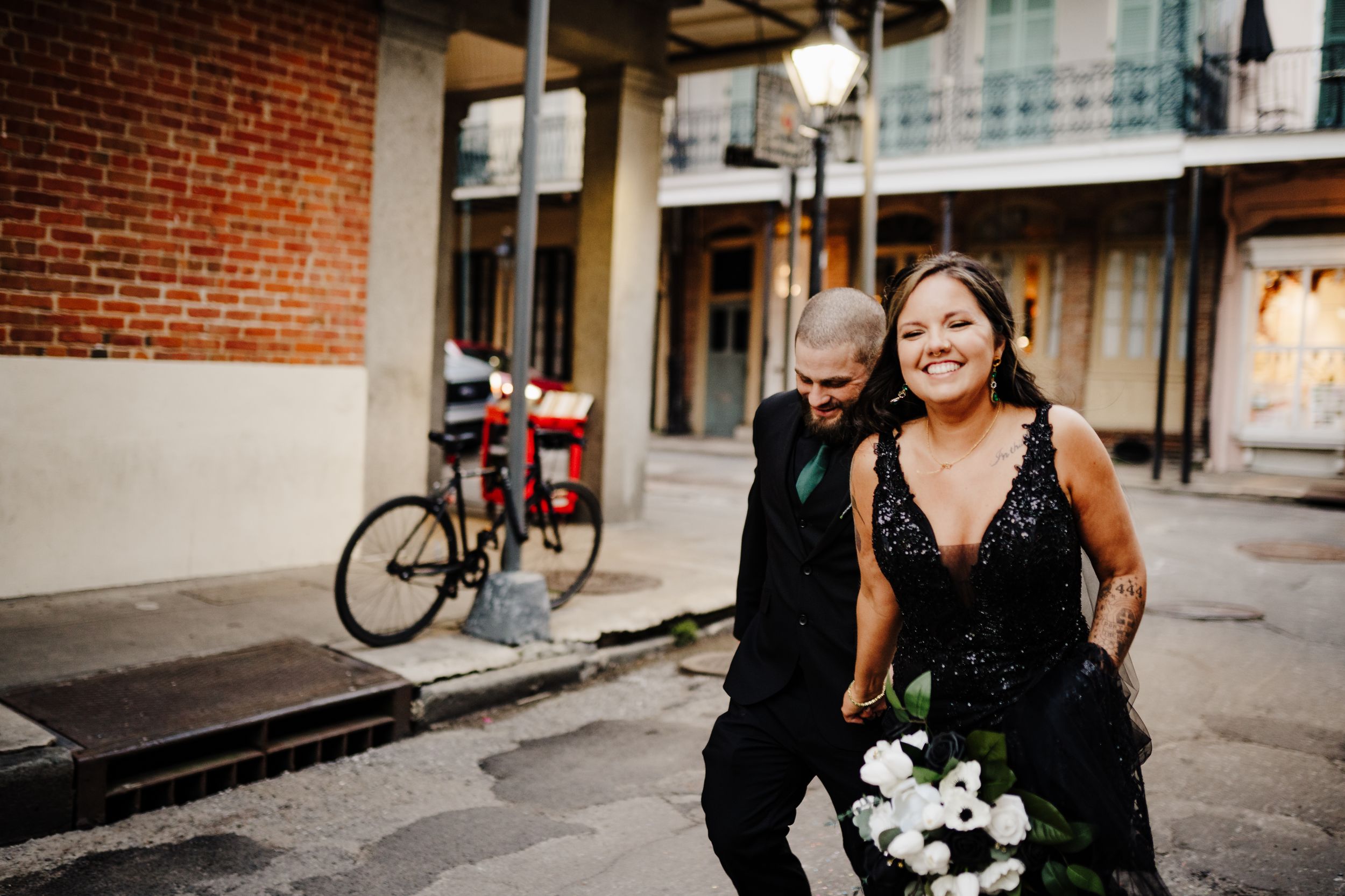 Two people walk down a city street with brick buildings and a bicycle in the background, one holding white flowers.