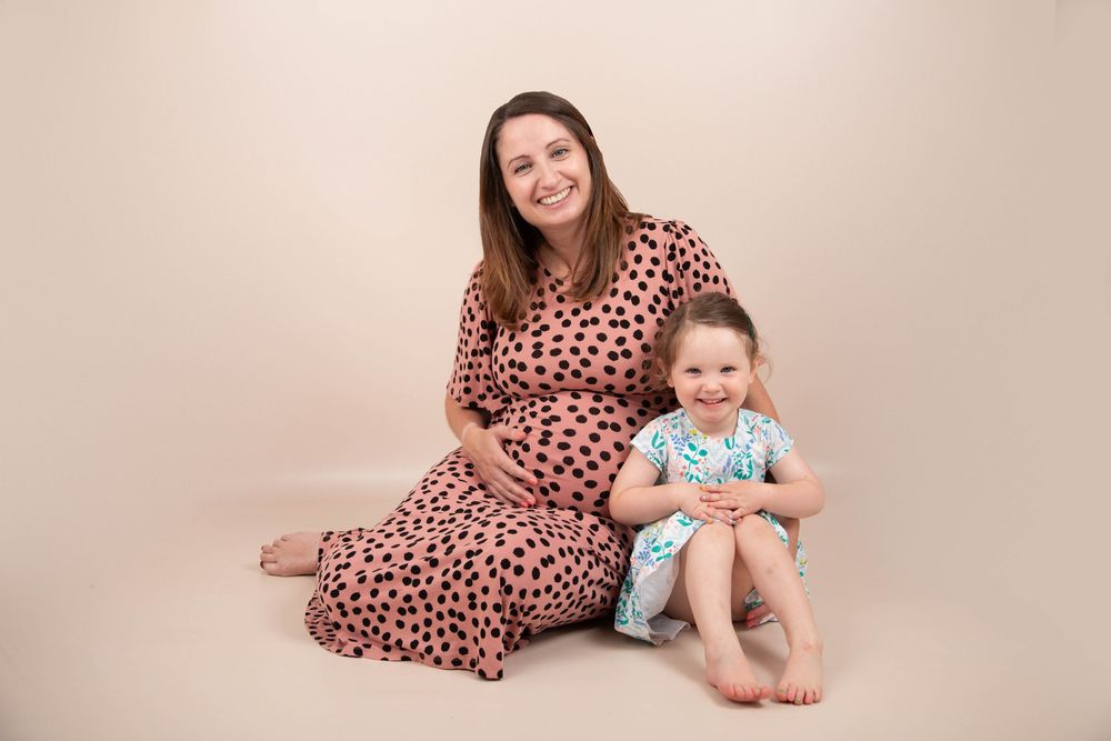 Smiling pregnant woman in pink polka dot dress sitting with young daughter on studio floor.