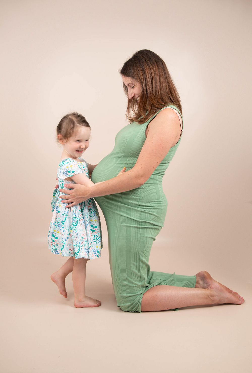 Pregnant woman in green dress kneeling as happy toddler daughter hugs her baby bump.