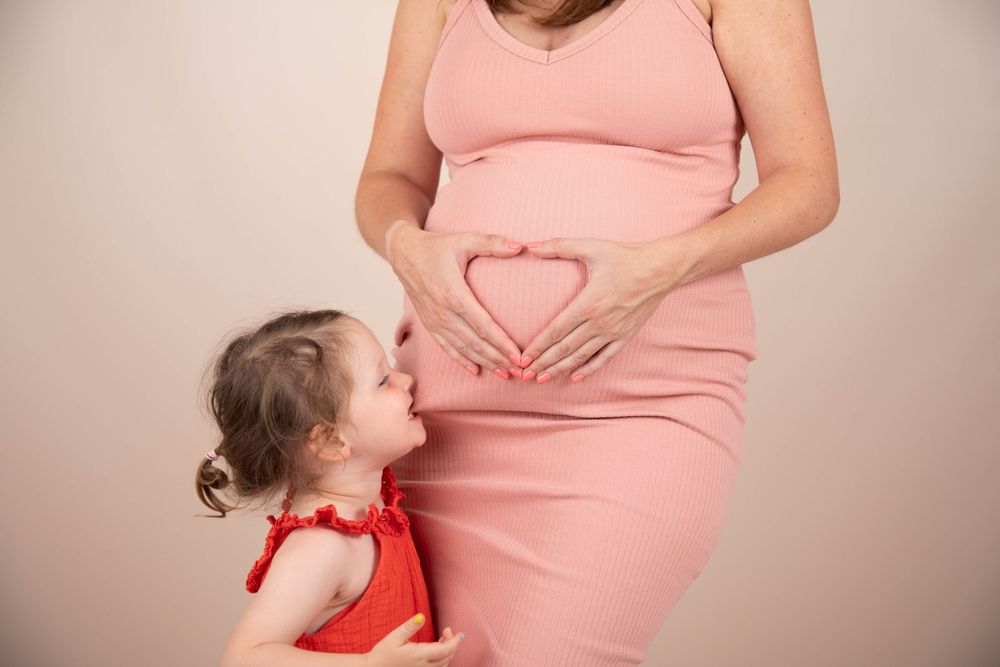 Pregnant woman in pink dress making heart shape on belly while toddler girl in red dress kisses bump.