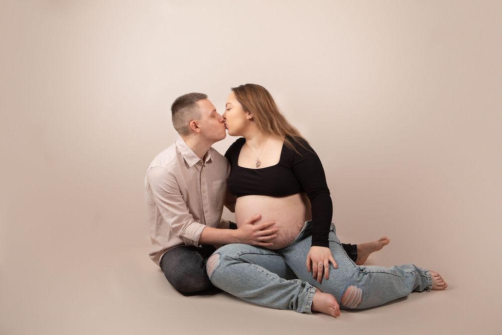 Couple sitting on floor kissing during maternity photoshoot, woman in black crop top and jeans.