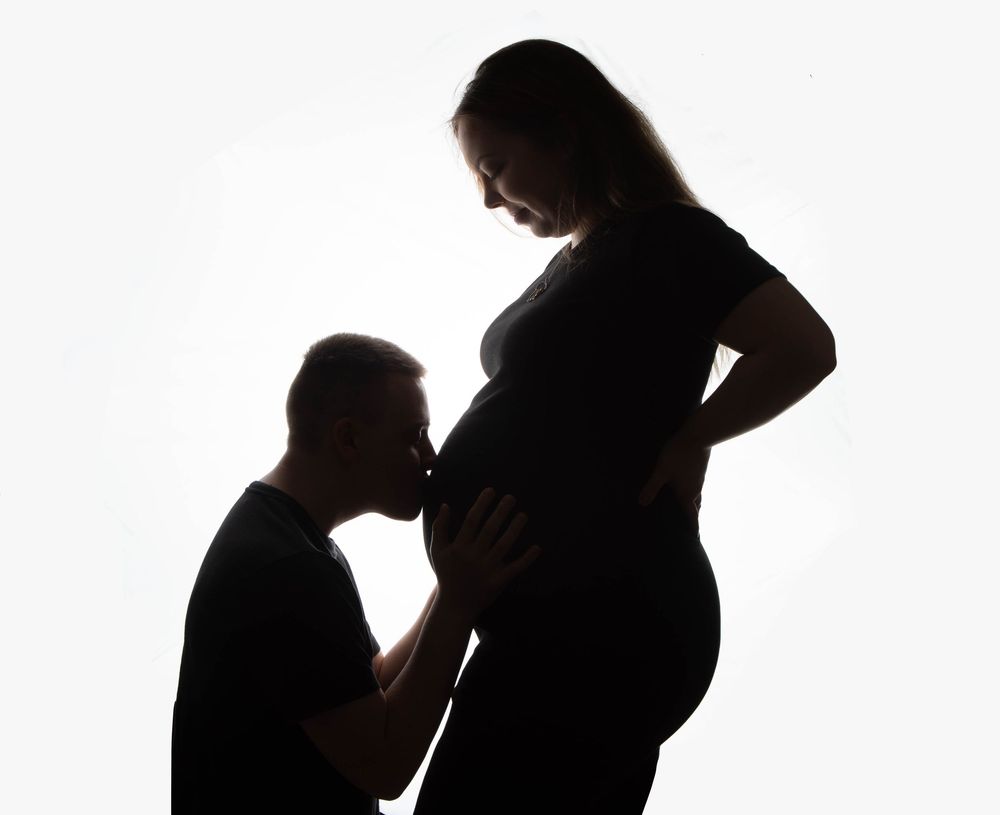 Silhouette of man kissing pregnant woman's baby bump against white background.