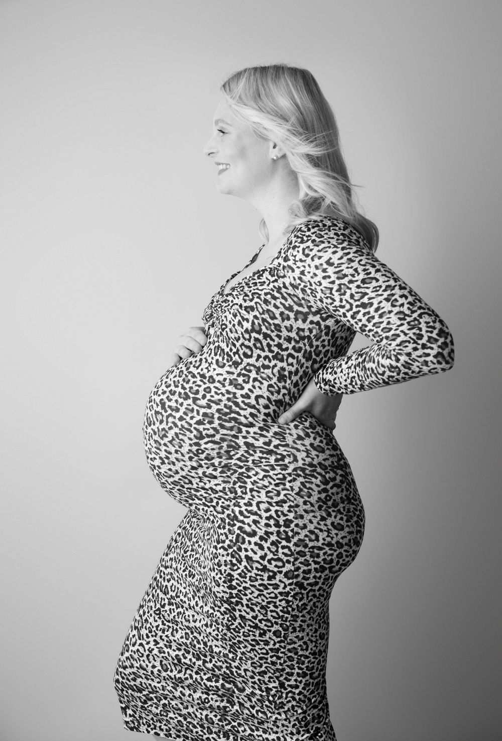 Black and white profile shot of smiling pregnant woman in leopard print dress holding bump.