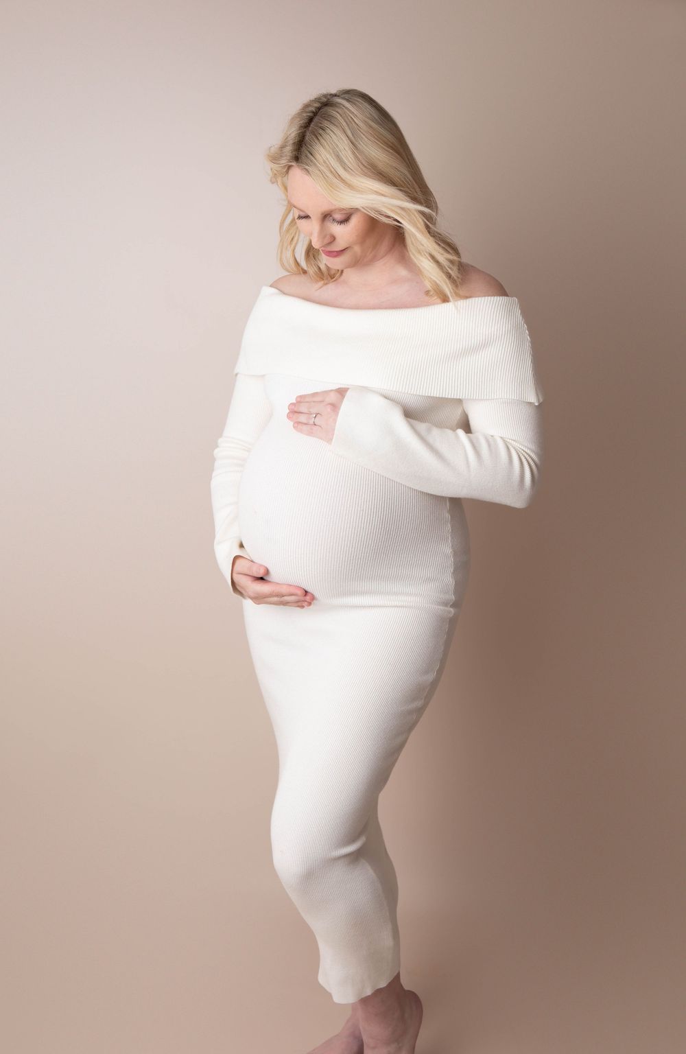 Pregnant woman in white off-shoulder dress cradling baby bump in studio maternity photo.