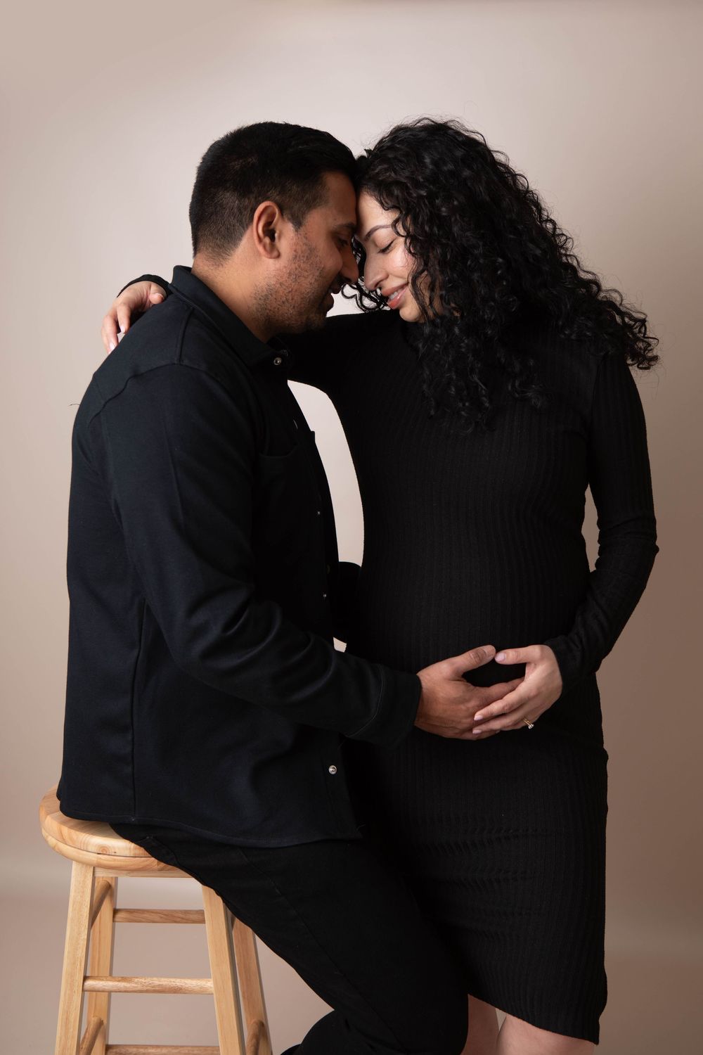 Couple in matching black outfits sharing tender moment during studio maternity photoshoot.