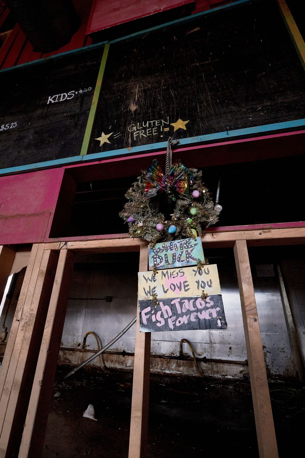 Damaged restaurant exterior with Christmas wreath and memorial signs reading We Miss You, We Love You, Fish Tacos Forever.