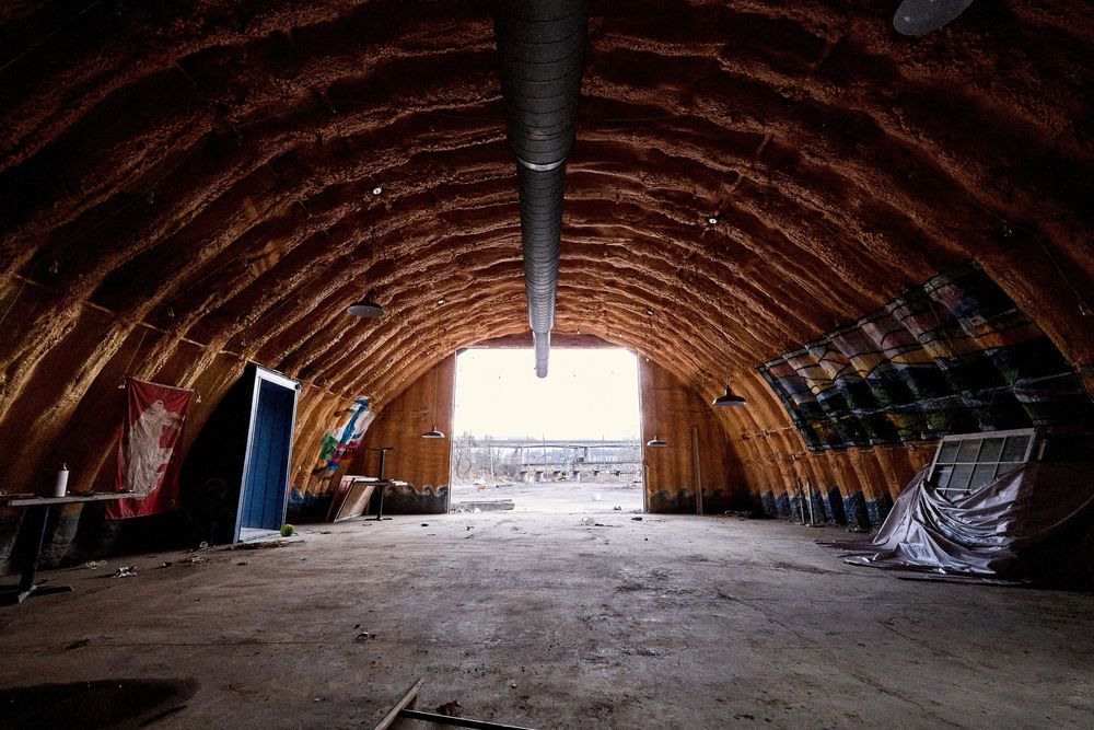 Interior of an abandoned Quonset hut with arched wooden ribbed ceiling and open end revealing daylight.