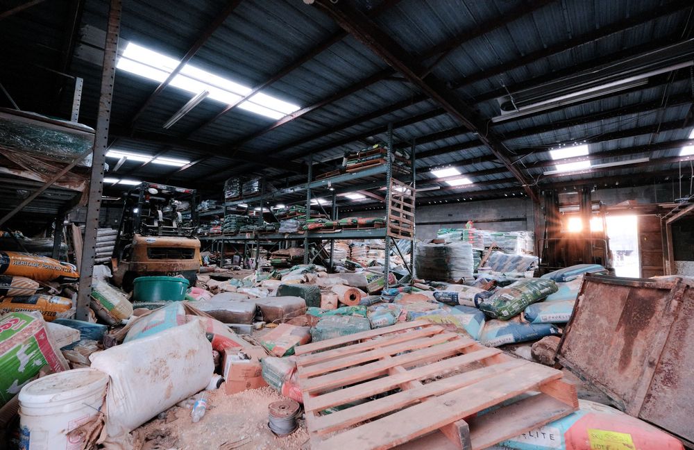Cluttered industrial warehouse interior filled with discarded materials, pallets, and debris under a metal roof.