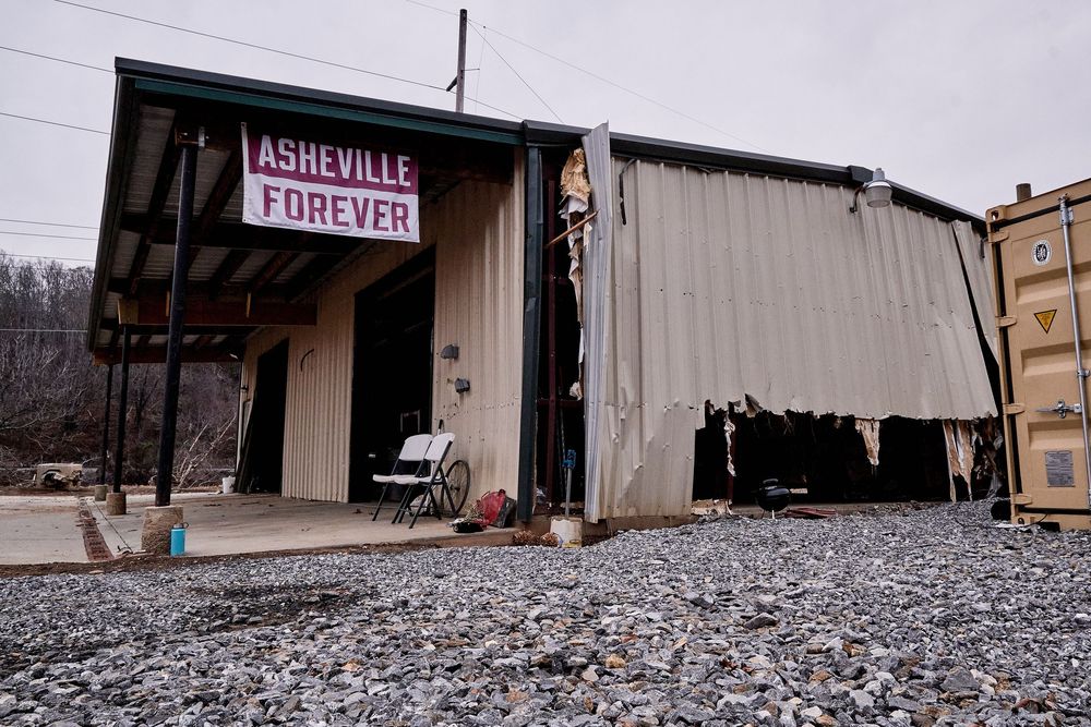 Damaged metal warehouse building with Asheville Forever banner on gravel lot after storm disaster.