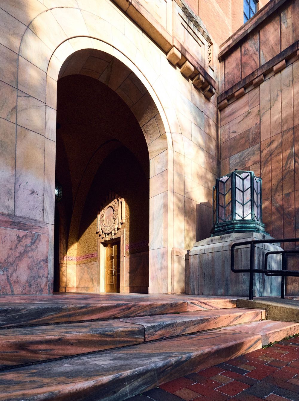 Grand stone archway entrance with marble steps and Art Deco lamp post casting warm sunlight shadows on ornate building facade.