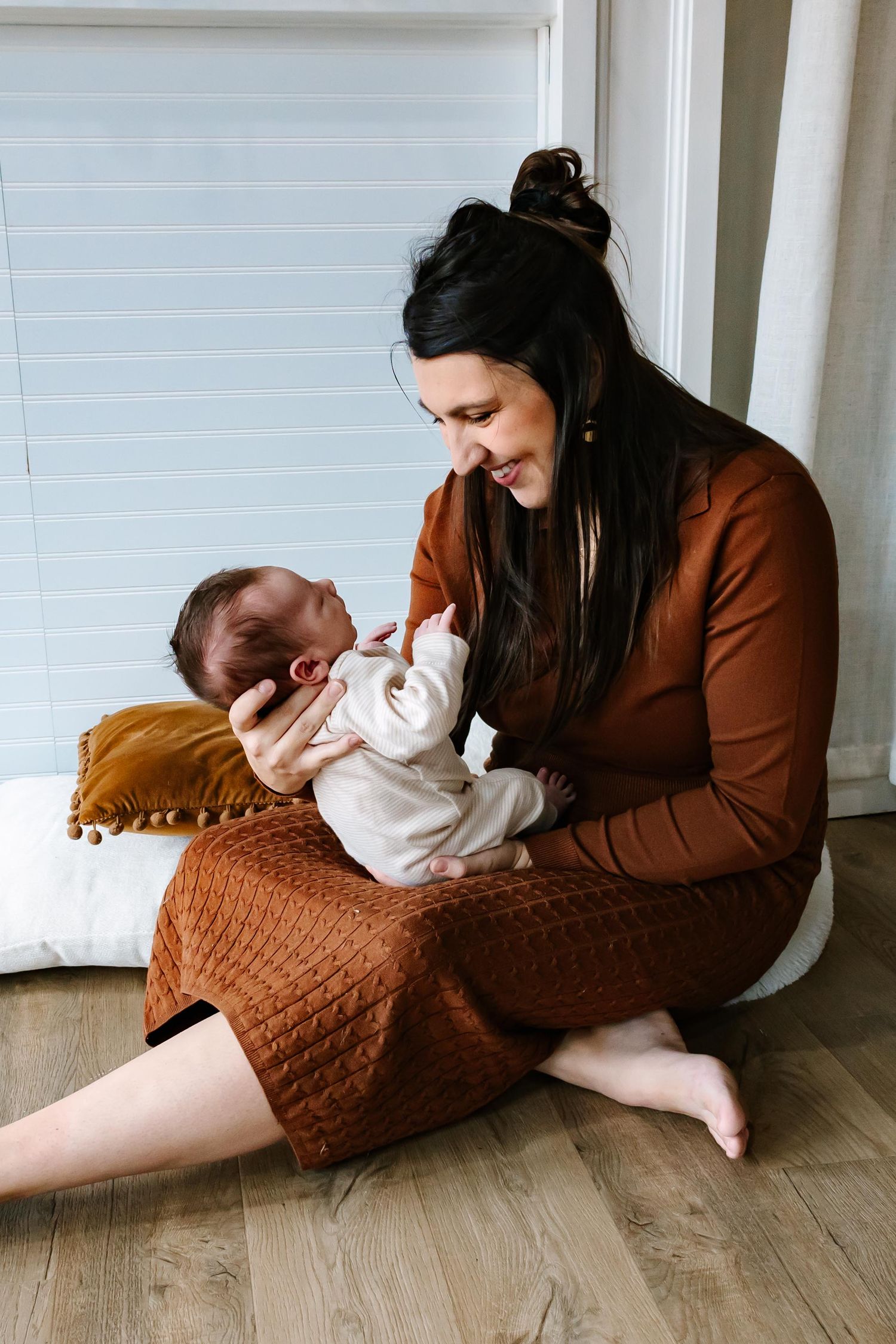 A mother in a brown dress sits on a wooden floor holding and gazing lovingly at her newborn baby wrapped in white.