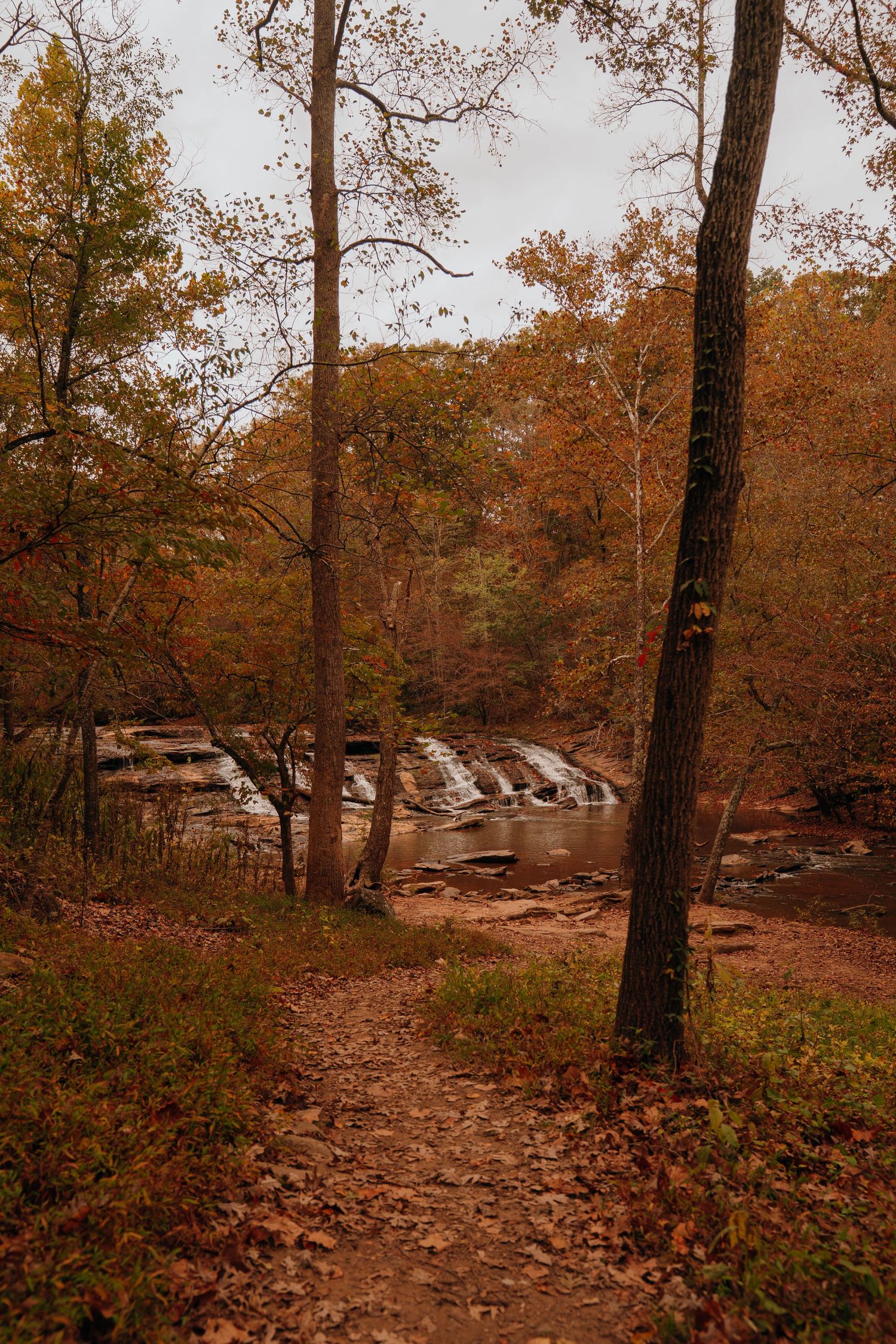 A dirt trail leads through autumn forest to a cascading waterfall with orange fall foliage reflecting in the water.