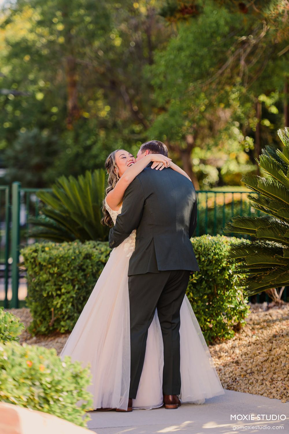 A couple embraces in a romantic moment on a garden path surrounded by lush greenery and palm plants.