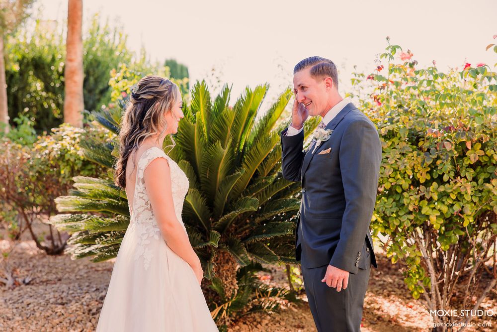 A couple shares an intimate moment in a lush garden setting with palm trees and flowering bushes in the background.