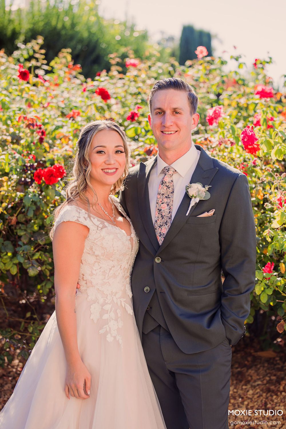 Wedding couple poses together in a beautiful rose garden with vibrant red blooms during sunset.