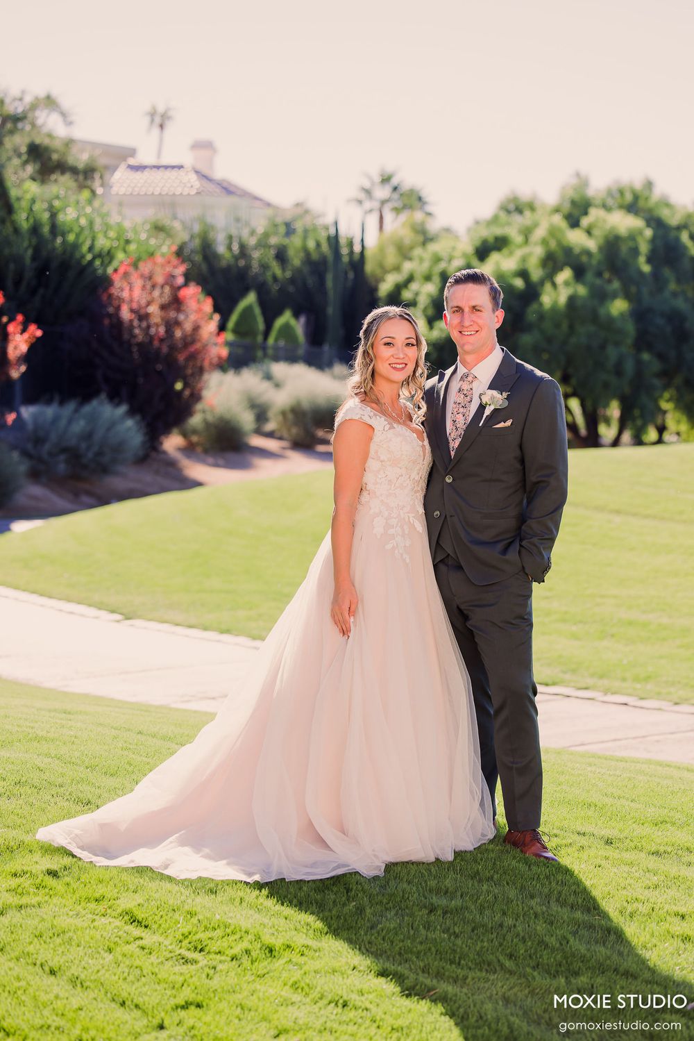 Newlyweds share romantic moments on a lush green lawn with garden scenery in the background.