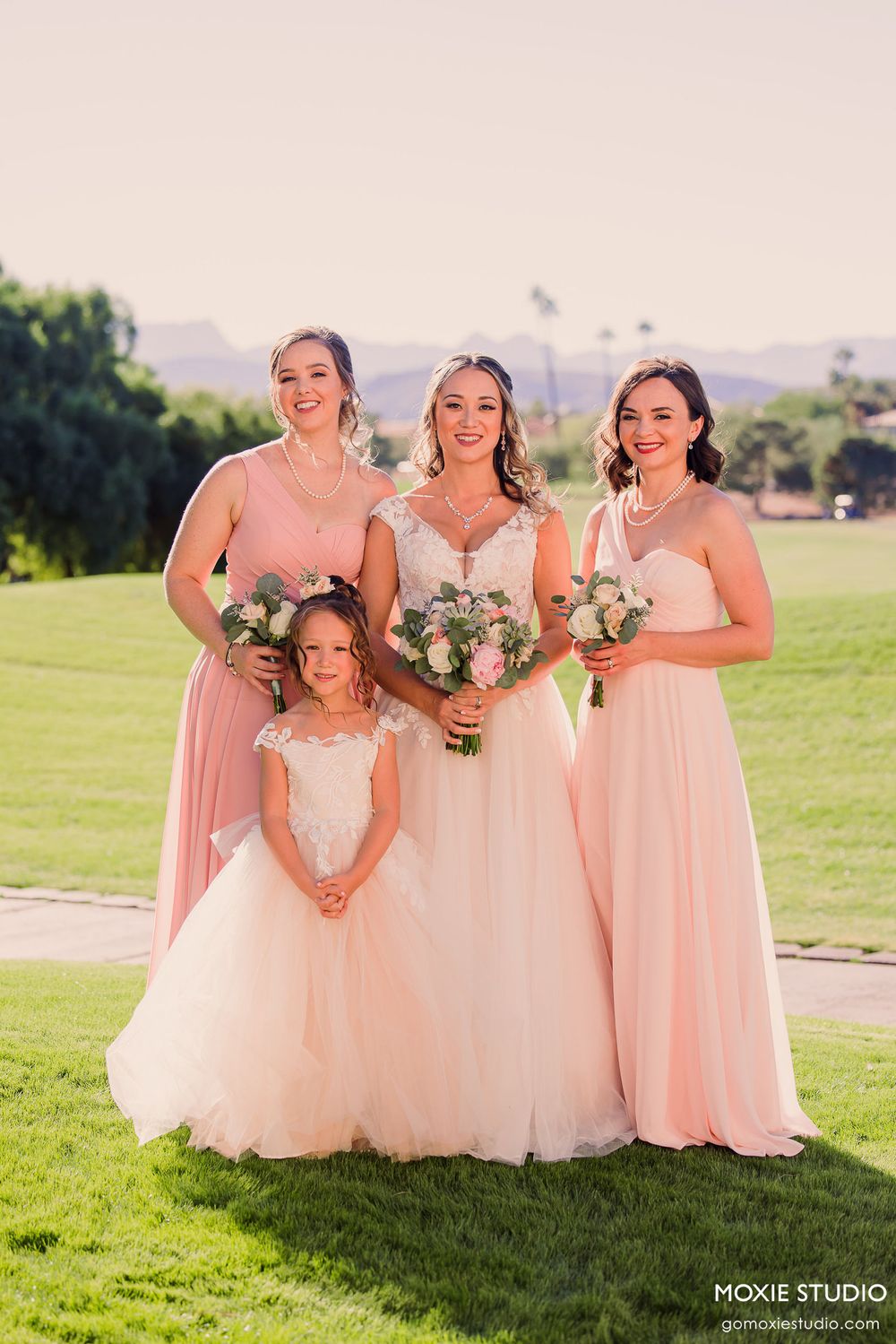 Bridesmaids in flowing peach gowns pose playfully on a sunny green lawn during an outdoor wedding photoshoot.