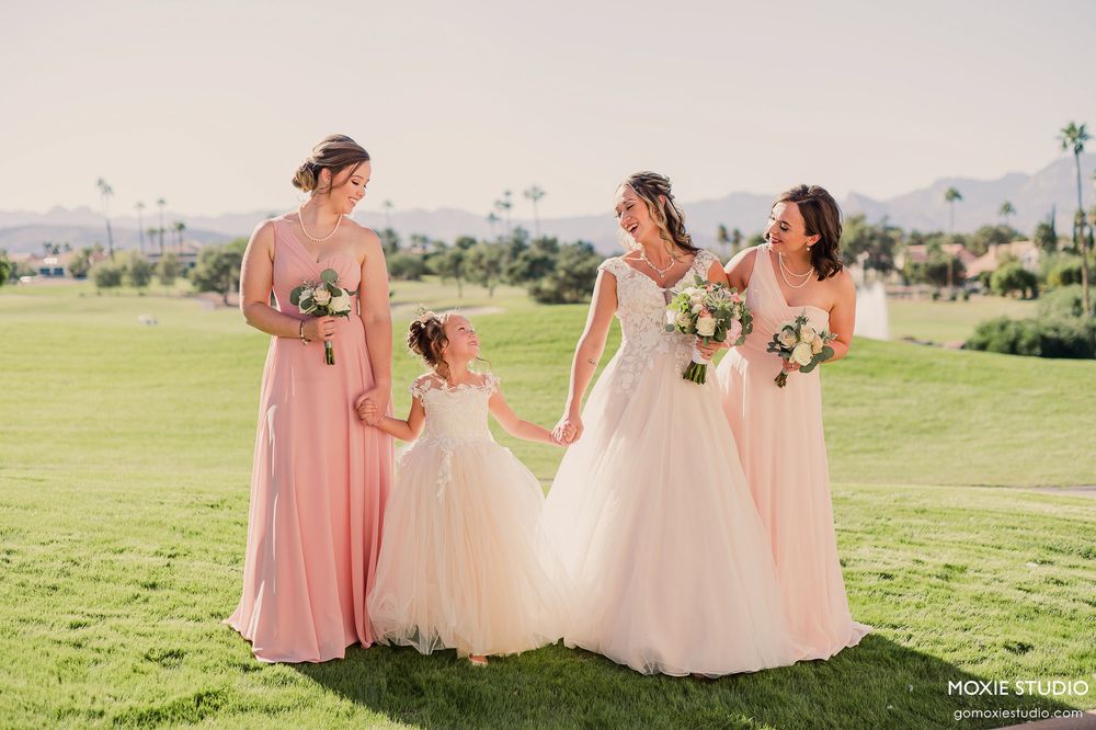 Bridesmaids in pink dresses and a flower girl in white pose on a sunny golf course with bouquets.
