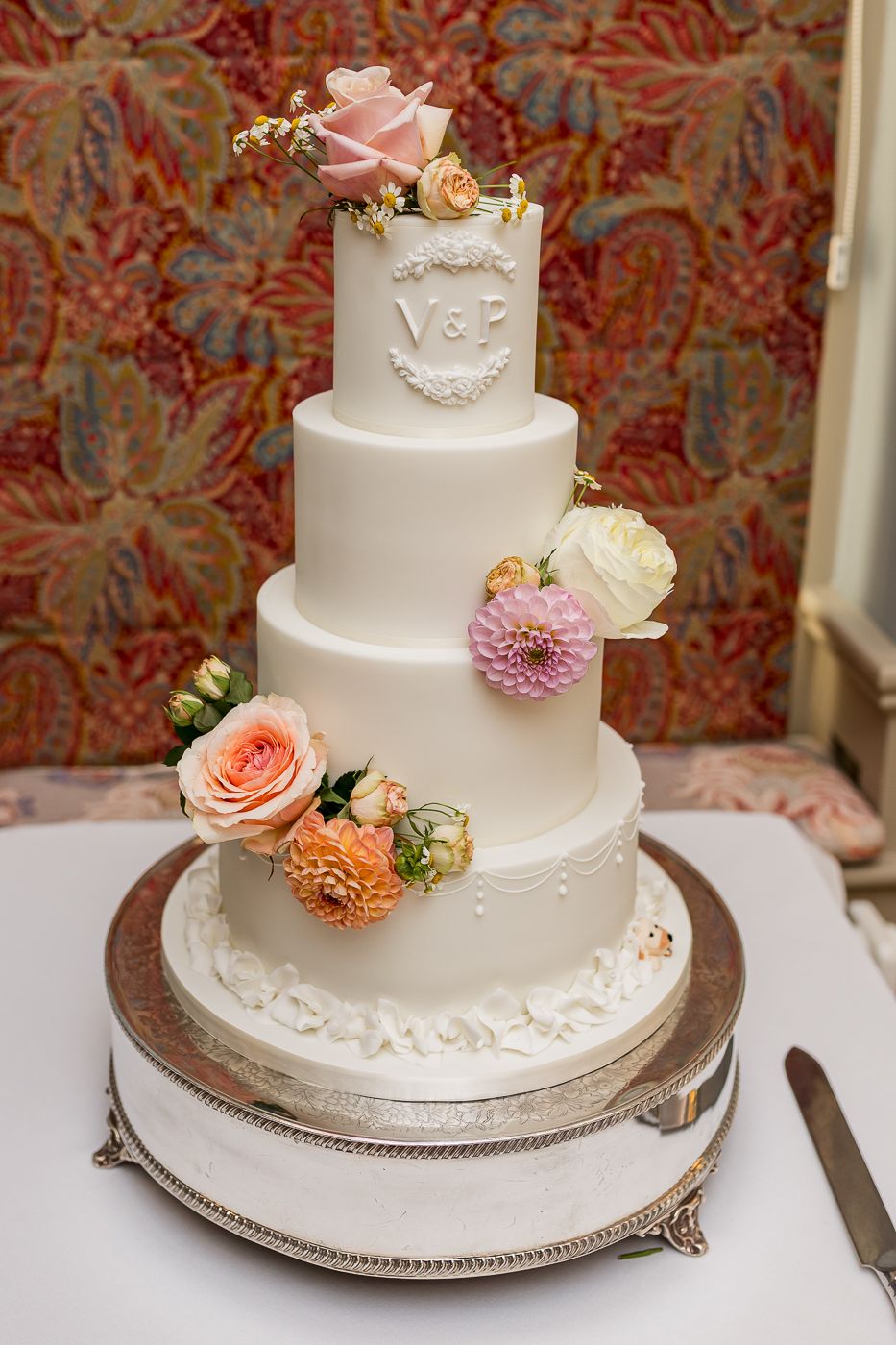 Elegant white wedding cake decorated with fresh pink and peach flowers on silver platter.