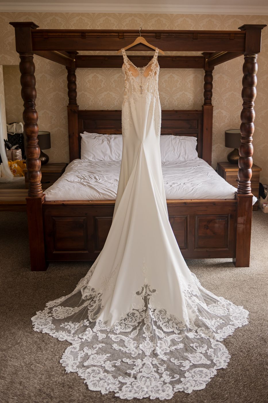 Wedding dress with intricate lace train displayed on wooden four-poster bed.