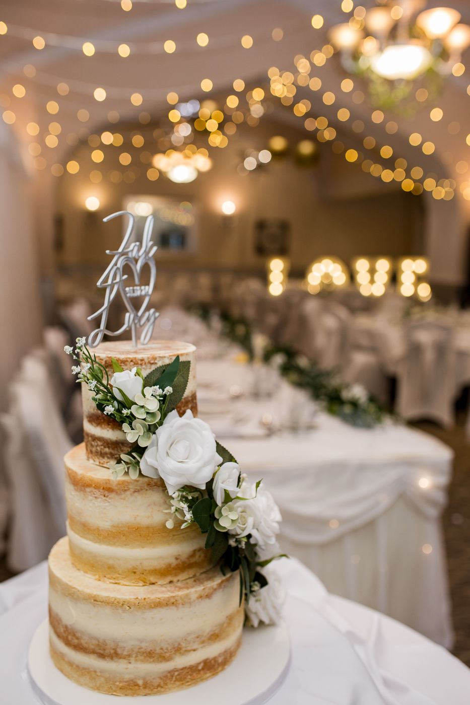 Rustic naked wedding cake decorated with white flowers and greenery on white tablecloth.