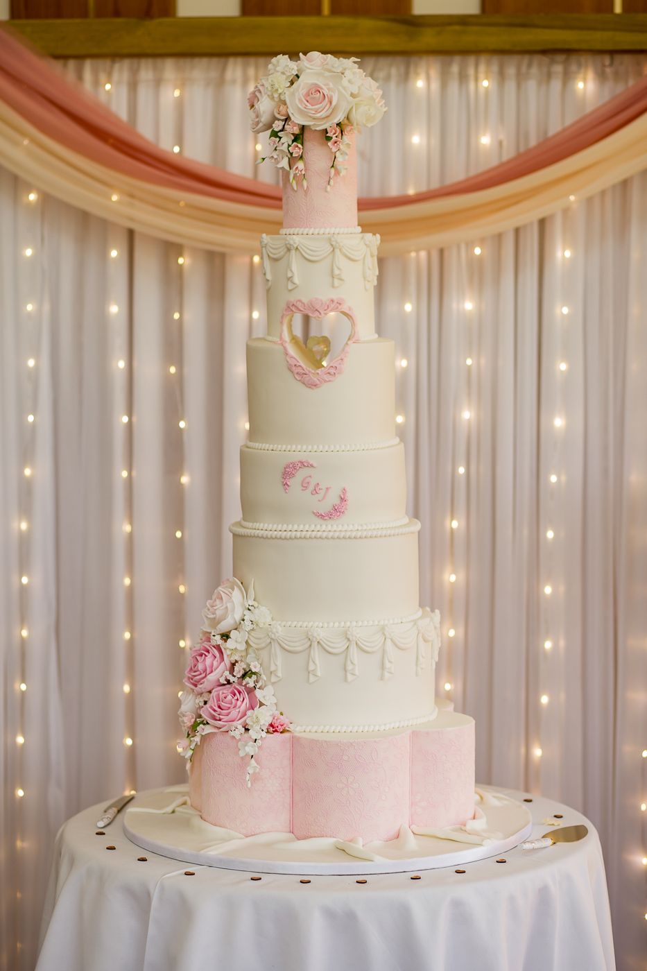 Elegant white and pink wedding cake with delicate floral decorations against a fairy light backdrop.