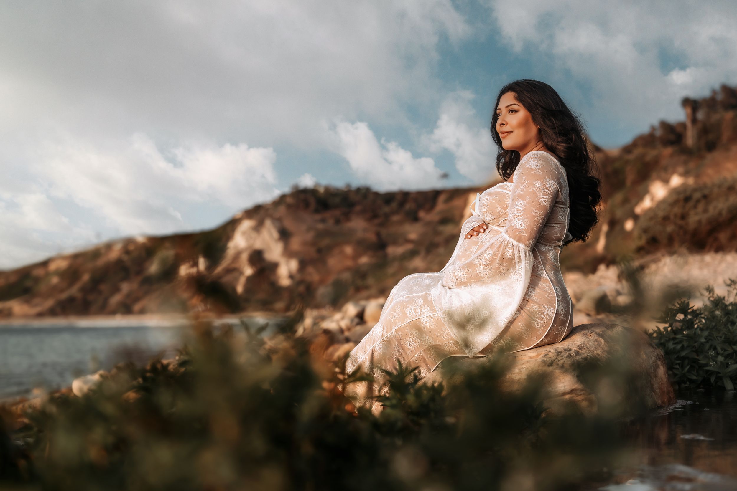 Woman in white dress posed elegantly on rocky coastal cliffs at sunset with dramatic cloudy sky in background.
