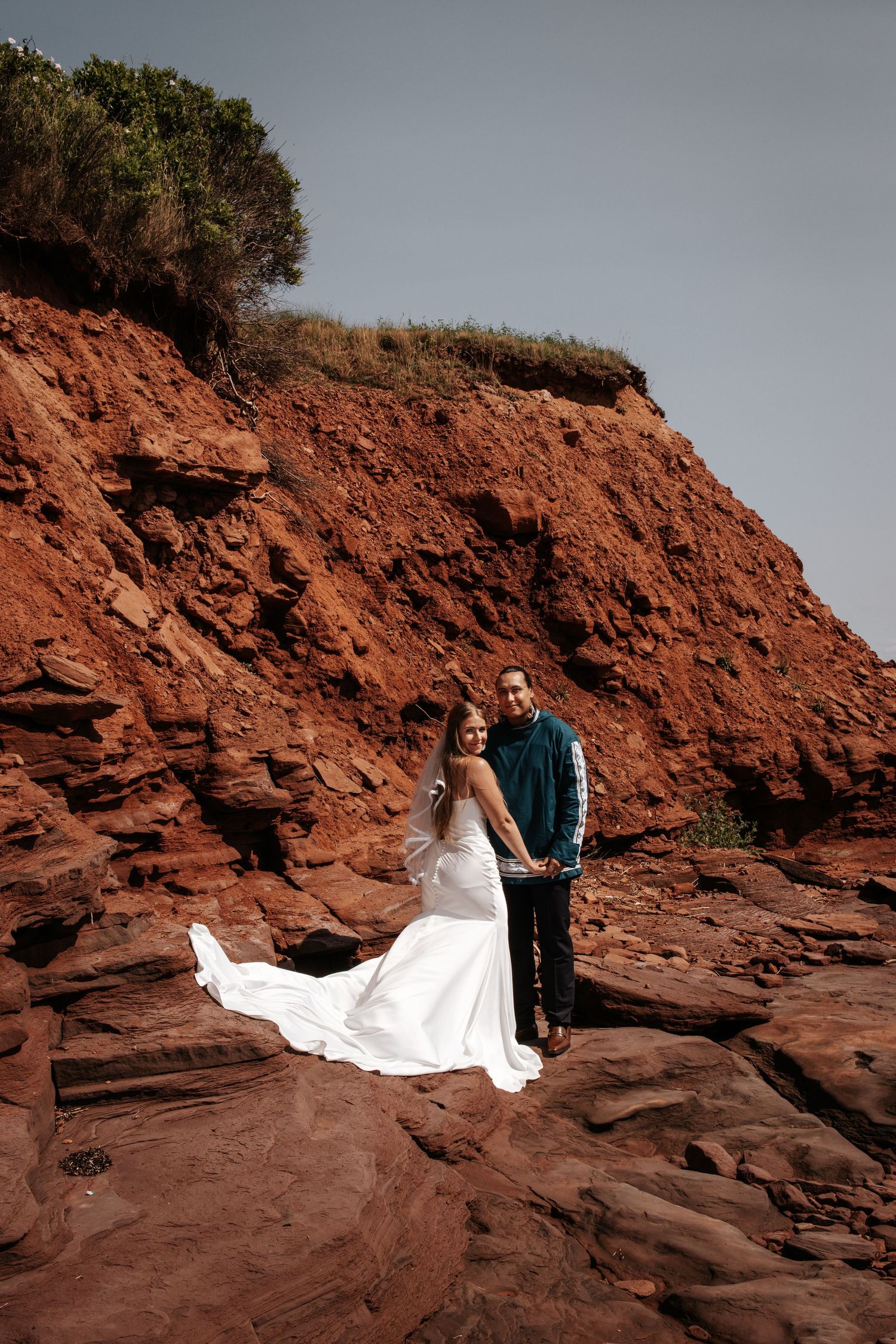 A wedding couple stands on dramatic red cliffside rocks with ocean views at sunset. Photo at Charlottetown PEI