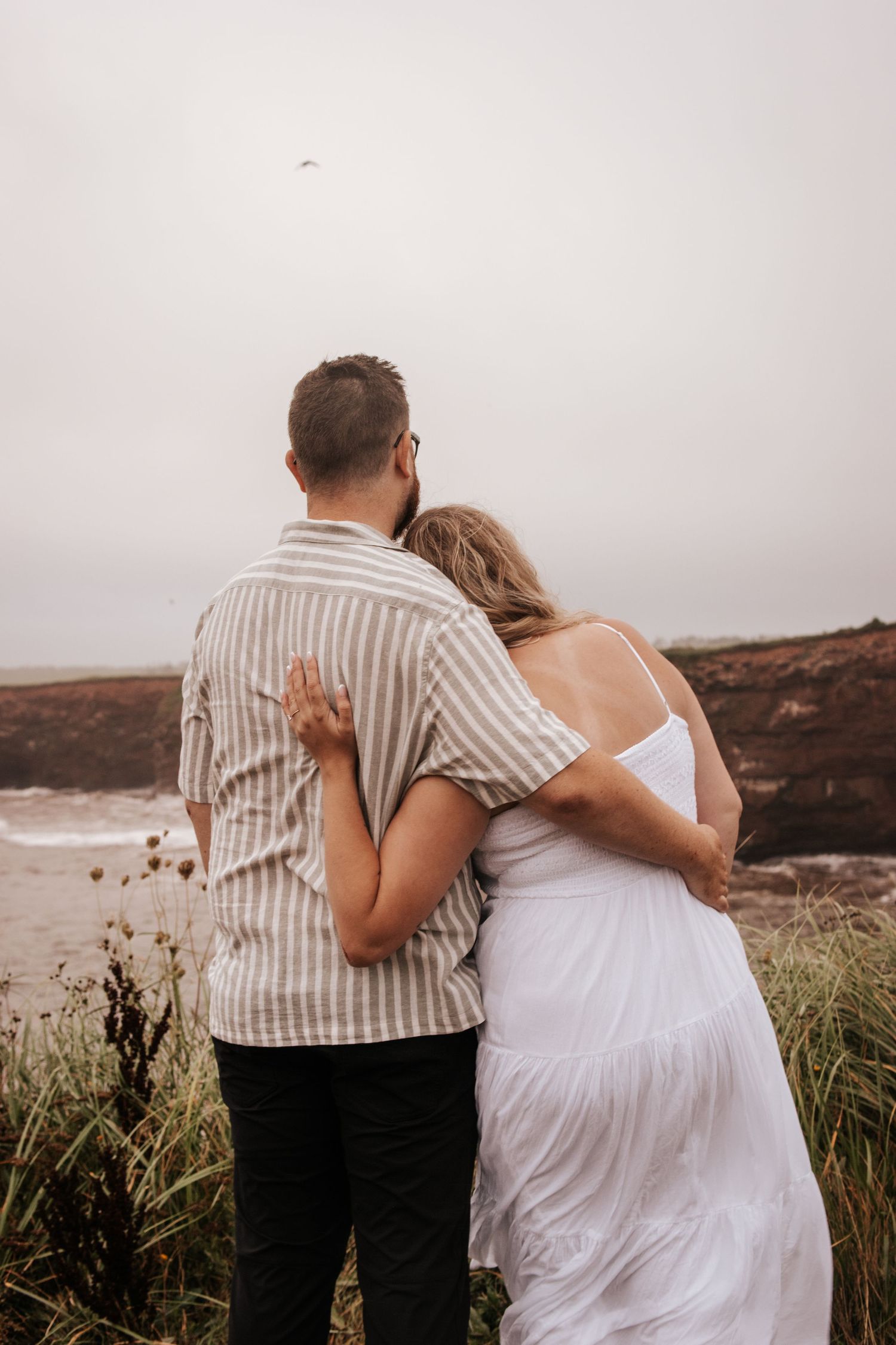 A couple embraces while looking out at the ocean from a cliff. Photo at Charlottetown PE