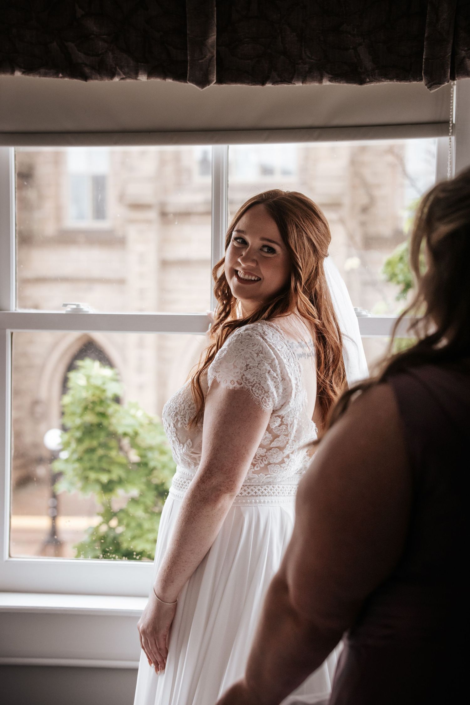 A bride in a lace wedding gown smiles by a window in natural light. Photo of St Dunstan's Basilica PEI