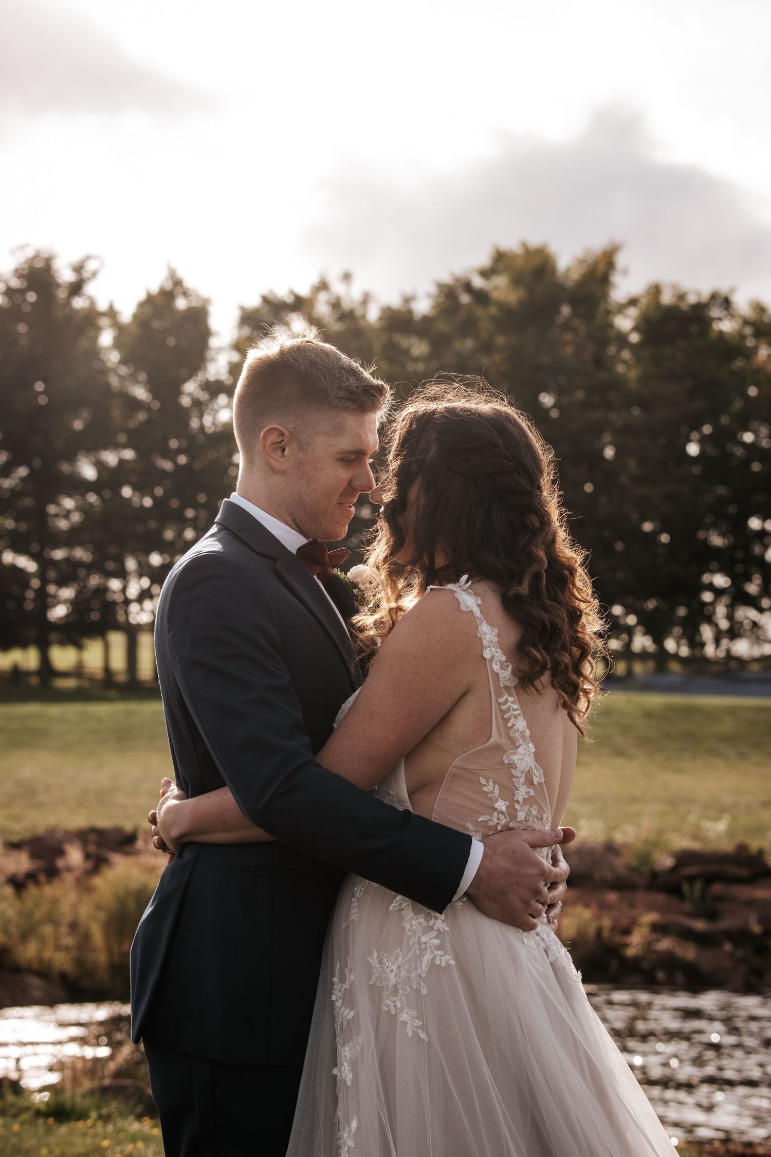 A romantic sunset wedding photo with a couple embracing in front of a tranquil pond and pine trees. Photo at Clinton Hills PEI