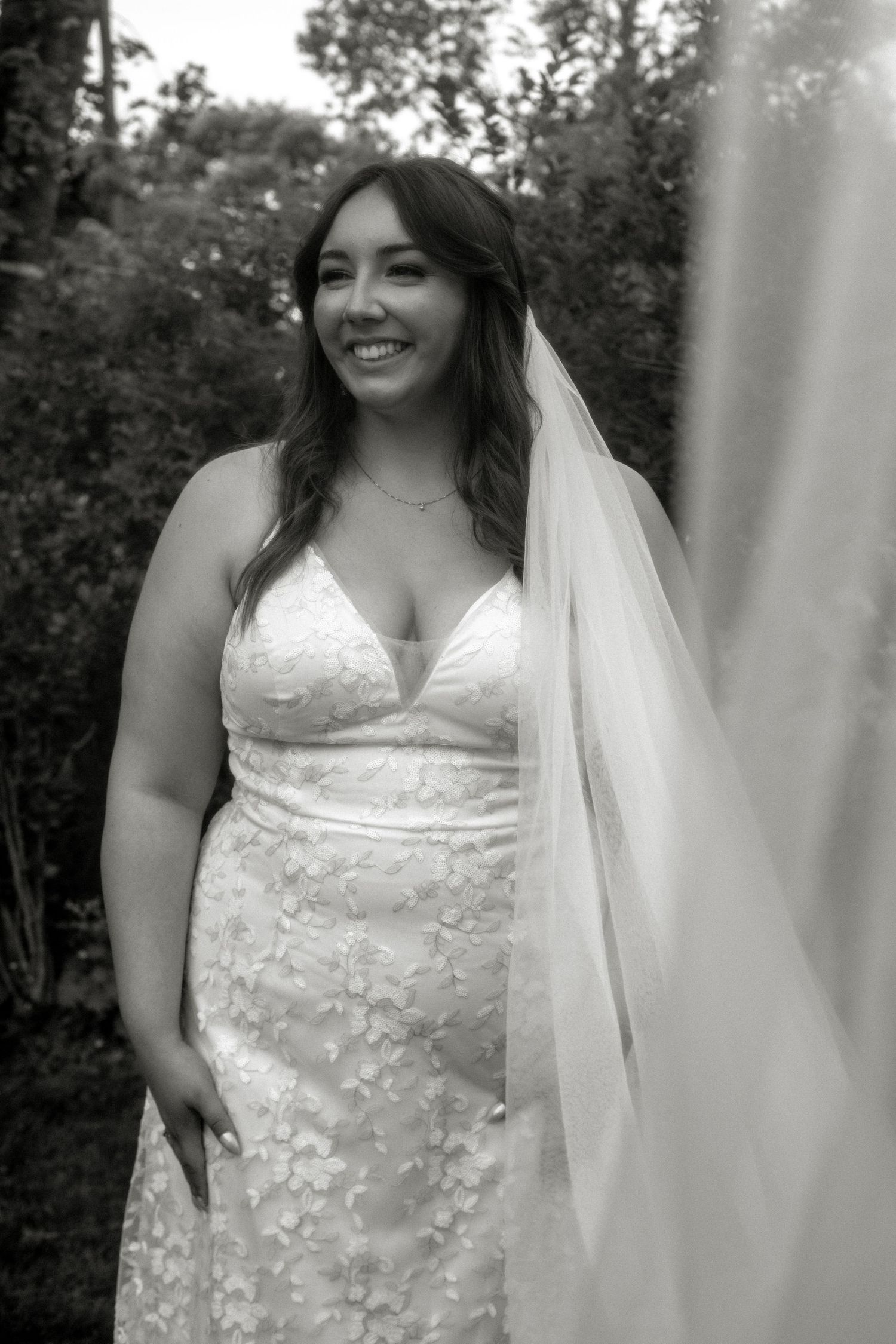 A bride in a lace wedding dress and veil smiles radiantly in a black and white portrait. Photo at Montague PEI