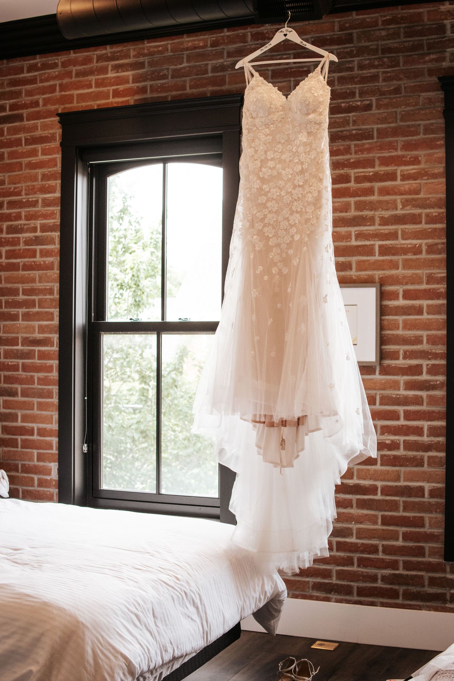 A delicate lace wedding dress hangs by a window in a rustic brick-walled room. Photo at Charlottetown PEI